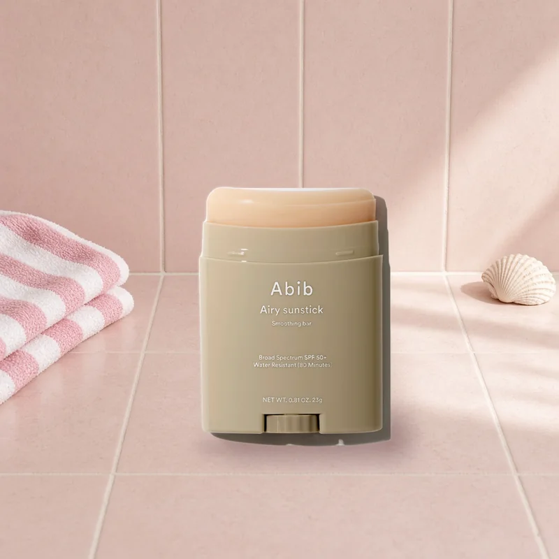 The Abib Airy Sunstick Smoothing Bar SPF50+ on a sunny blush tiled surface beside a striped beach towel and a seashell