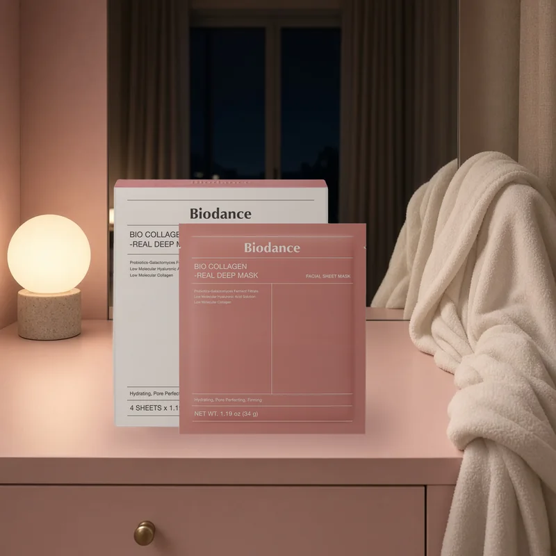 The BIODANCE Bio-Collagen Real Deep Mask box and sheet mask pouch on a blush vanity at night beside a glowing lamp and a white robe