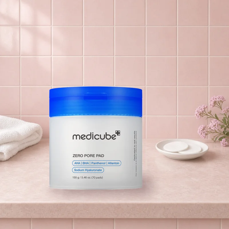 The medicube Zero Pore Pad jar with a blue lid on a blush tiled vanity beside a towel and a dish of toner pads