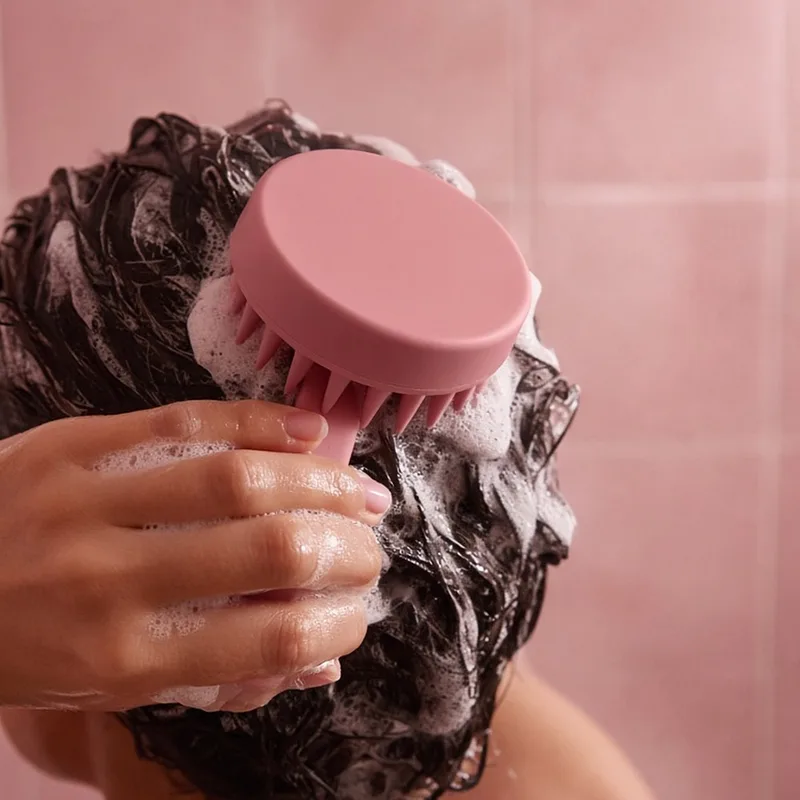 Illustration of a hand using a pink silicone scalp massager brush in lathered dark hair, standing in for the HEETA brush